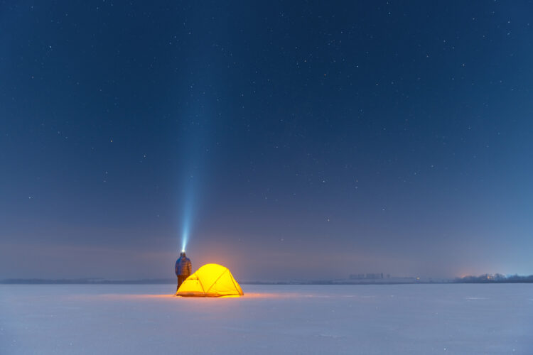 Tourist with flashlight near yellow tent lighted from the inside against the backdrop of incredible starry sky. Amazing night landscape. Tourists camp in snowy field. Travel concept